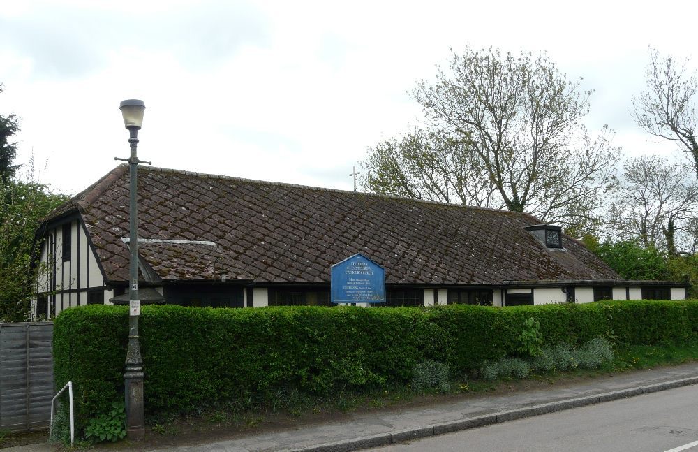 St Thomas of Canterbury church: a long, low black-and-white building with a steep tiled roof and a small cross, behind a hedge, with its blue noticeboard